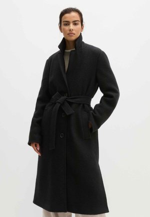 Woman wearing a long black belted coat with a high collar, standing with one hand in pocket against a plain light background.