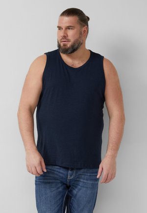 Brodaty mężczyzna z włosami związanymi do tyłu, ubrany w granatową koszulkę bez rękawów i niebieskie dżinsy, stoi na tle jednolitej jasnej ściany.