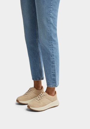 Beige suède sneakers met een gevoerde zool en vetersluiting. Gecombineerd met lichtblauwe, taps toelopende jeans, wat een casual outfit laat zien.