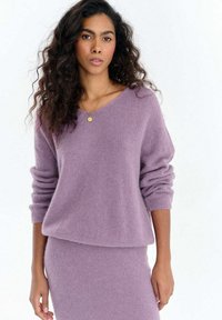 Fioletowy, miękki sweter dzianinowy z dekoltem w serek i długimi, lekko bufkowatymi rękawami. Noszony na dopasowaną spódnicę w tym samym kolorze. Tkanina swetra wygląda na puszystą.