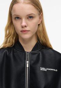 Νεαρή γυναίκα με μακριά ξανθά μαλλιά, φορώντας ένα μαύρο δερμάτινο μπουφάν με το λογότυπο "Karl Lagerfeld Jeans" στο στήθος, κοιτάζοντας μπροστά σε λευκό φόντο.