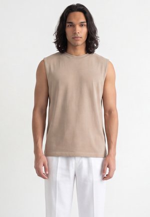 Uomo con capelli fino alle spalle che indossa una camicia beige senza maniche e pantaloni bianchi, in piedi davanti a uno sfondo chiaro e semplice.