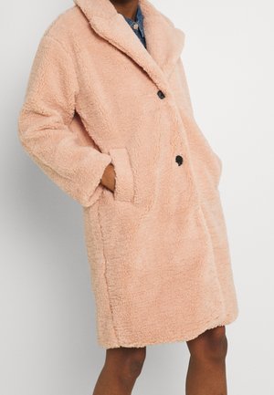 Persoon draagt een teddyjas van pastelroze, knie lang met brede rever, zwarte knopen en handen in de zijzakken gestopt.