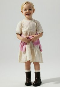 Robe beige clair à manches courtes et au design en étages. Pull rose noué à la taille. Bottines marron et chaussettes crème.