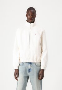 Hvid lysjakke med høj krave og lynlås foran, med elastiske manchetter og et lille logo på venstre bryst. Båret med blå jeans.