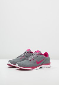 Zapatillas deportivas grises con acentos rosas, parte superior de malla ligera, collar acolchado y suela blanca. Presenta un prominente logo de Nike en color rosa.