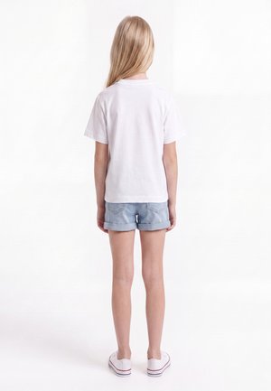 Niño con cabello rubio largo de espaldas lleva una camiseta blanca lisa, pantalones cortos de mezclilla azul claro remangados y zapatillas blancas.