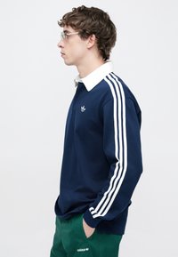 Μπλε ναυτικό πόλο με μακριά μανίκια, λευκό γιακά και τρεις λευκές ρίγες σε κάθε μανίκι. Διαθέτει μικρό κεντητό λογότυπο στο στήθος.