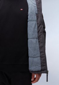 Foret jakke med en mørkegrå ydre skal, lys grå fleeceforing og en sort sweatshirt under, med synlig lynlås og manchet detalje.