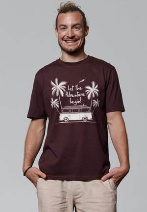 Bordeaux t-shirt met een witte grafische weergave van een vintage camperbus met palmbomen en de tekst "Laat het avontuur beginnen!" in een speels lettertype.