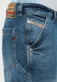 Κοντινή λήψη μπλε τζιν Diesel Industry που δείχνει την πίσω τσέπη, την πλαϊνή τσέπη, τις θηλιές της ζώνης και το δερμάτινο λογότυπο στη μέση.