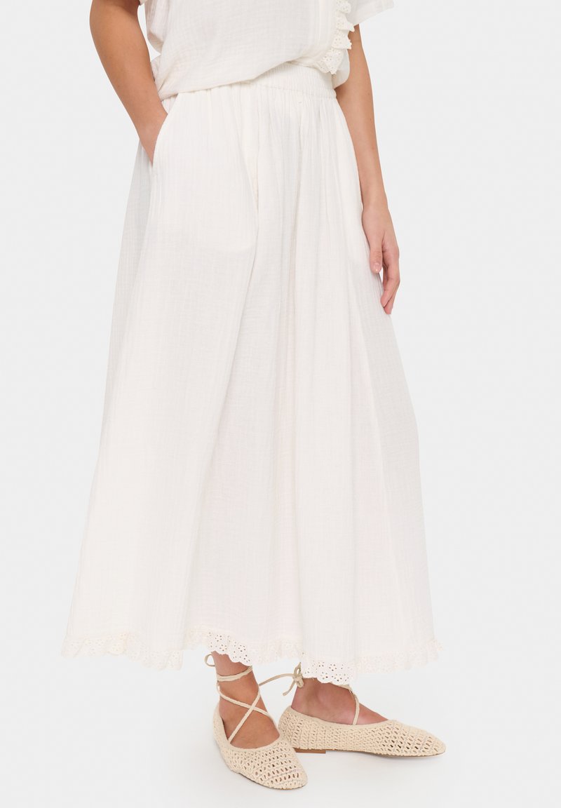 Jupe maxi blanche avec taille élastique, tissu texturé, détail de dentelle à bord festonné. Assortie à des ballerines beiges en tissu tissé avec des lacets à la cheville.