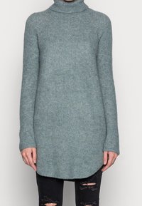 Světlý tyrkysový svetr s vysokým límcem a dlouhými rukávy, s žebrovanou texturou a zakřiveným lemem, nošený přes černé roztrhané džíny.