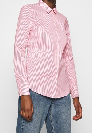 Personne portant une chemise ajustée à manches longues à rayures verticales roses et blanches, boutonnée, associée à un jean bleu, debout devant un fond uni.