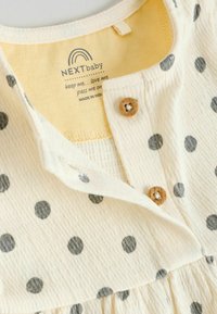 Robe pour bébé couleur crème avec des pois gris, dotée d'une fermeture par boutons et d'une texture de tissu douce. L'étiquette indique la marque et les instructions d'entretien.