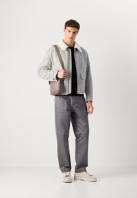 Tânăr îmbrăcat cu o jachetă gri cu carouri și guler alb, pantaloni gri, adidași bej și purtând o geantă de umăr mauve, stând pe un fundal alb.