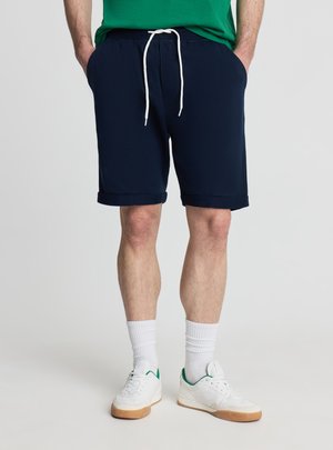 Mężczyzna ubrany w granatowe szorty z podwiniętymi brzegami i białym sznurkiem, białe skarpetki do kostek oraz białe trampki z zielonymi akcentami.