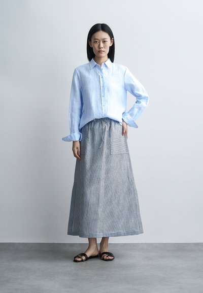 Femme debout contre un mur uni, portant une chemise bleu clair à manches longues, une jupe rayée longue jusqu'aux chevilles avec une poche et des sandales noires.