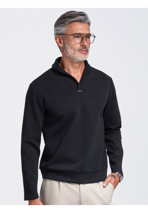 Mand med gråt hår og skæg iført briller, sort pullover, beige bukser og ur, står med den ene hånd i lommen.