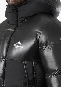 Veste noire matelassée avec col montant et capuche, en tissu brillant, design matelassé, avec un logo Helvetica blanc sur la poitrine.