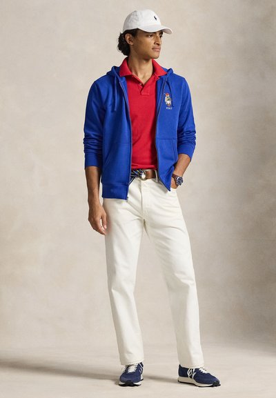 Modrá mikina na zip, červené polo tričko, bílé kalhoty a tmavě modré tenisky. Mikina má vyšívané logo a pásek s kovovou sponou.