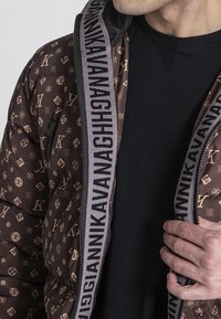 Hnedá zateplená bunda s textúrovaným vzorom log, so zipsovým uzáverom a výrazným čiernym lemom s logom pozdĺž prednej časti a goliera.