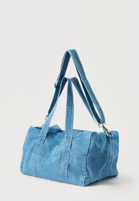 Μπλε τσάντα duffel από τζιν με δύο κοντά χερούλια και ρυθμιζόμενο, αποσπώμενο ιμάντα ώμου σε ανοιχτό φόντο.