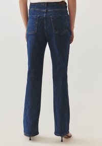 Jeans acampanados de denim azul oscuro con cinco bolsillos, costuras visibles y cintura de altura media. Textura suave con un ligero desgaste cerca del dobladillo.