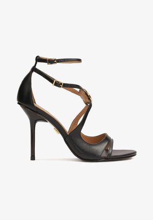 Black high-heeled sandal with a sleek leather design, featuring crisscross straps, an ankle buckle, and a smooth, pointed toe. Gold accents visible.