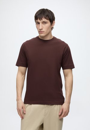 Joven con el pelo corto y oscuro, lleva una camisa marrón lisa de manga corta y pantalones beige, de pie frente a un fondo claro y liso.