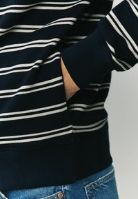 Sötét navy csíkos pulóver, fehér vízszintes csíkokkal, bordázott mandzsettával és elülső zsebbel, kék farmernadrággal párosítva.