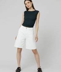 Jeune femme se tenant les jambes écartées, portant un haut sans manches noir, un short blanc jusqu'aux genoux, des sandales noires et des boucles d'oreilles créoles, sur fond gris.