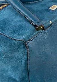 Gros plan sur le coin d’un sac en cuir et daim bleu avec des bords cousus et un fermoir en métal doré qui relie la poignée.