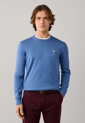 Junger Mann mit blauem Langarm-Pullover und bordeauxroter Hose steht mit einer Hand in der Tasche vor einem einfarbig grauen Hintergrund.
