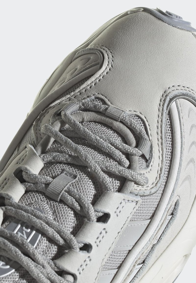 Hellgraue Sneaker aus Leder und Mesh, mit strukturierten Akzenten, runder Form und mehreren Schnürpunkten in sportlichem Design.