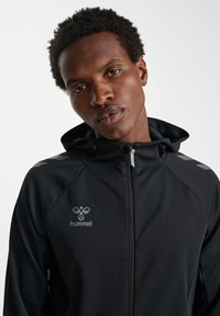 Zwarte gewatteerde jas met capuchon, gemaakt van een glad, lichtgewicht materiaal. Voorzien van een ritssluiting aan de voorkant, grijs logo en reflecterende accenten op de schouders.