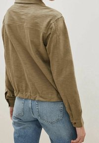 Chemise légère olive avec un col, des poignets boutonnés et un dos légèrement court, associée à un jean en denim bleu clair.
