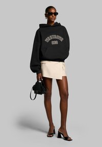 Black oversized hoodie with white lettering, beige wrap skirt, black handbag, and black heeled sandals. Sunglasses and hoop earrings worn.