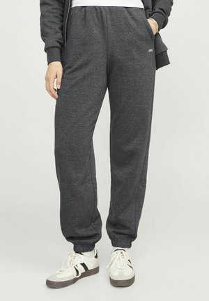 Persoon die donkergrijze joggingbroek met elastische boorden en witte sneakers met zwarte strepen draagt, staand met één hand in de zak.