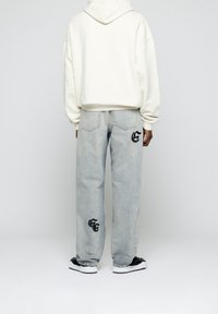 Persona con jeans a gamba larga blu chiaro con loghi neri cuciti sul retro e sulla parte inferiore della gamba, felpa bianca con cappuccio e sneakers in bianco e nero.