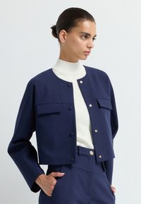 Granatowa krótka kurtka z dwoma przednimi kieszeniami, okrągłym dekoltem i złotymi guzikami, noszona na białym, teksturowanym swetrze z golfem.