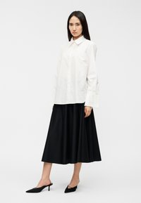 Λευκό πουκάμισο με γιακά και κουμπιά, συνδυασμένο με μαύρη, αέρινη μίντι φούστα. Μαύρα ψηλοτάκουνα παπούτσια με μυτερή μύτη ολοκληρώνουν το σύνολο. Λείο ύφασμα, μινιμαλιστικός σχεδιασμός.