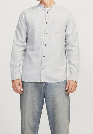 Homme portant une chemise gris clair à manches longues avec col mandarin et un jean bleu délavé, debout devant un fond uni.