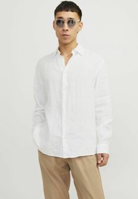 Weißes Leinenhemd mit langen Ärmeln, Button-Down-Kragen und lockerer Passform, kombiniert mit beigen Hosen. Glatte Textur, minimalistisches Design.