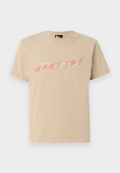 Beige cotton t-shirt featuring a graphic print with white text reading "THE WHAT IS?" and orange accents in a bold, slanted font.