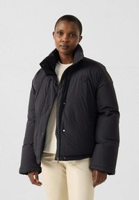 Zwarte donsjas met een hoge kraag en drukknopen, met een logo op de borst en zijzakken, gedragen over een licht shirt.