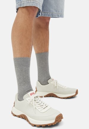 White sneakers with a textured surface, brown rubber outsole, and red logo. Paired with gray ribbed socks, worn with denim shorts.