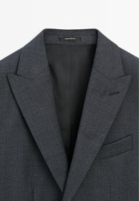 Grauer, strukturierter Blazer mit schmalem Revers, einer einzigen Brusttasche und glattem Innenfutter. Das Etikett trägt die Aufschrift "Made in Portugal".