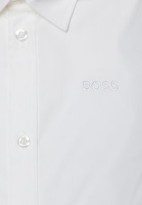 Balta marškiniai su mygtukais, pagaminti iš lygaus audinio, su smailiu apykakle, ant krūtinės turi subtiliai išsiuvinėtą "BOSS" logotipą, atitinkančios baltos spalvos.