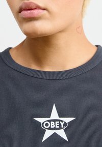 Mörkgrå ribbade t-shirt med en vit stjärndesign och texten "OBEY." Inkluderar en liten hjärt-tatuering på nackområdet.